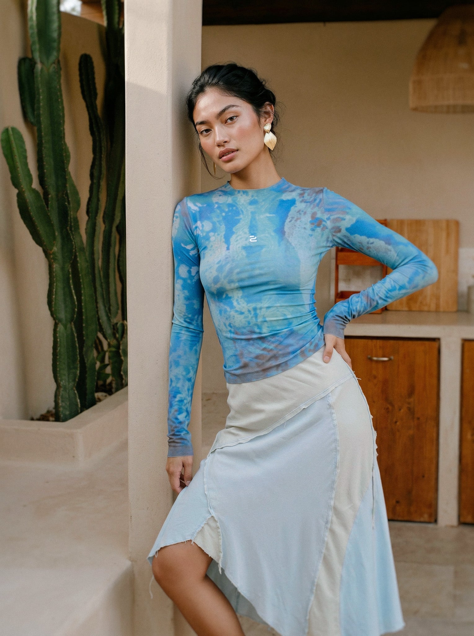 Woman wearing a blue floral top and white skirt standing in a casual setting with plants and furniture.
