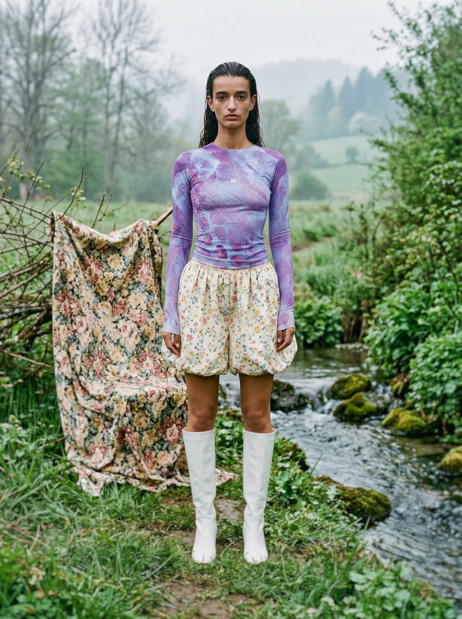 Woman standing in a natural setting wearing a purple top and white shorts with a floral garment draped over a tree branch.