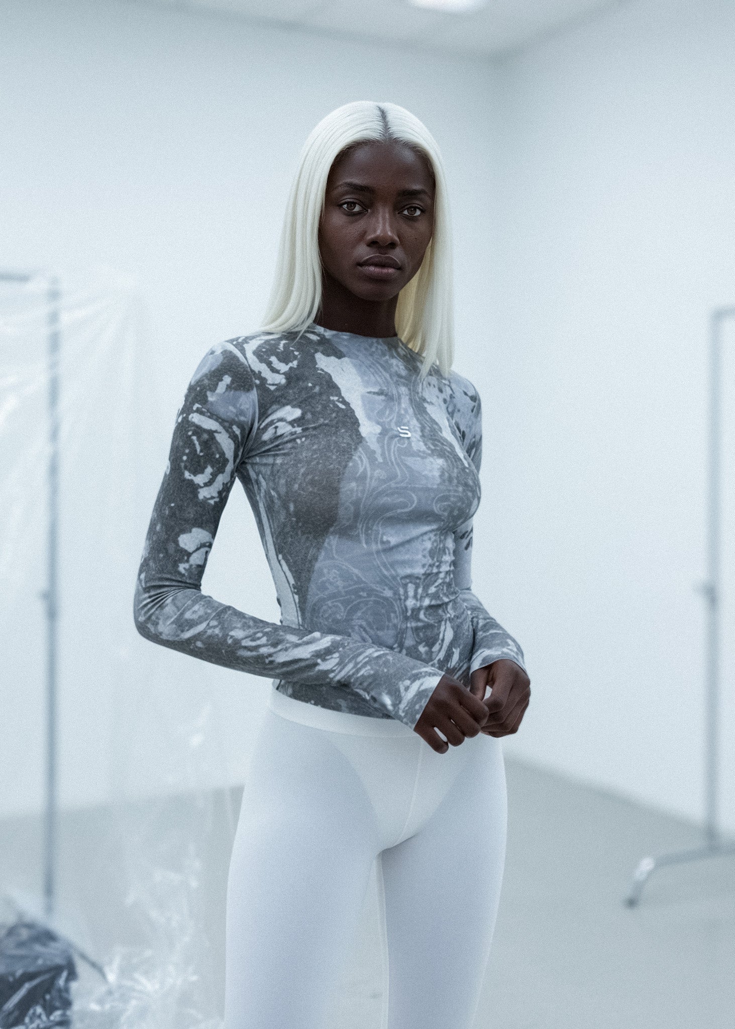 Person wearing a patterned long-sleeve top and white pants in a minimalistic setting