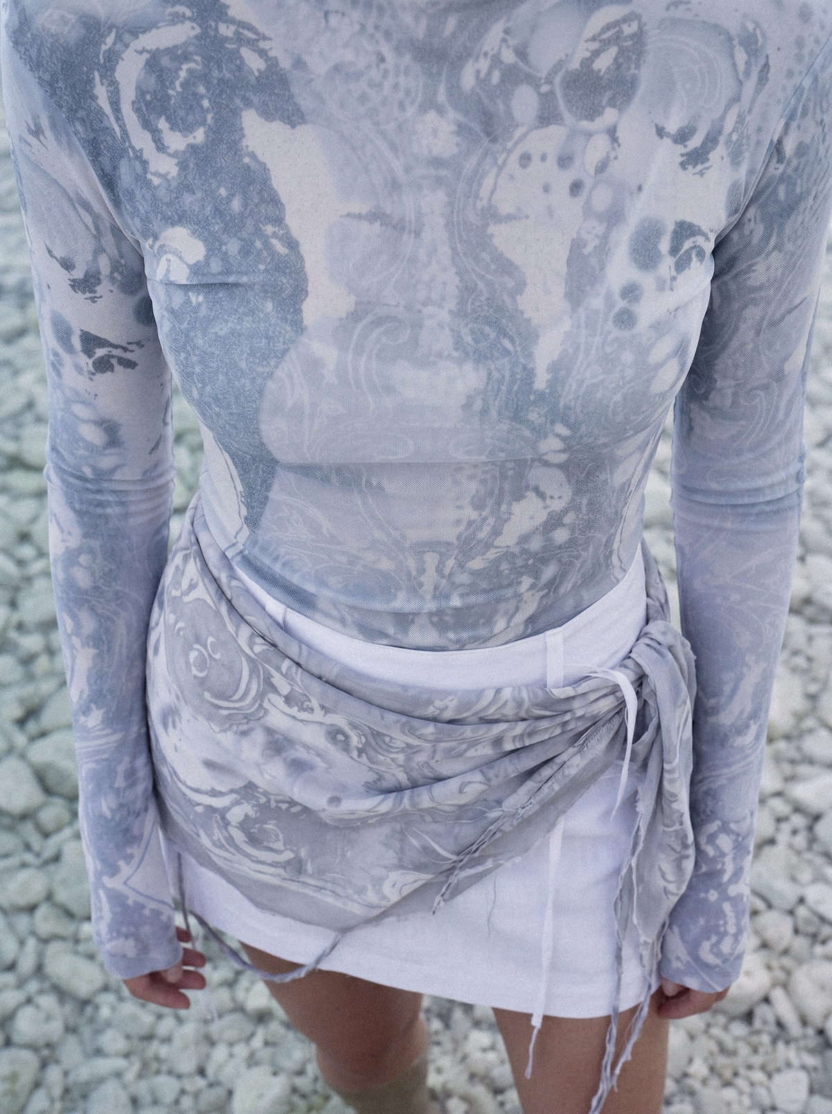 A person wearing a fitted long-sleeve top and matching wrap-style skirt in a light gray and white abstract print, standing outdoors on a pebble surface.