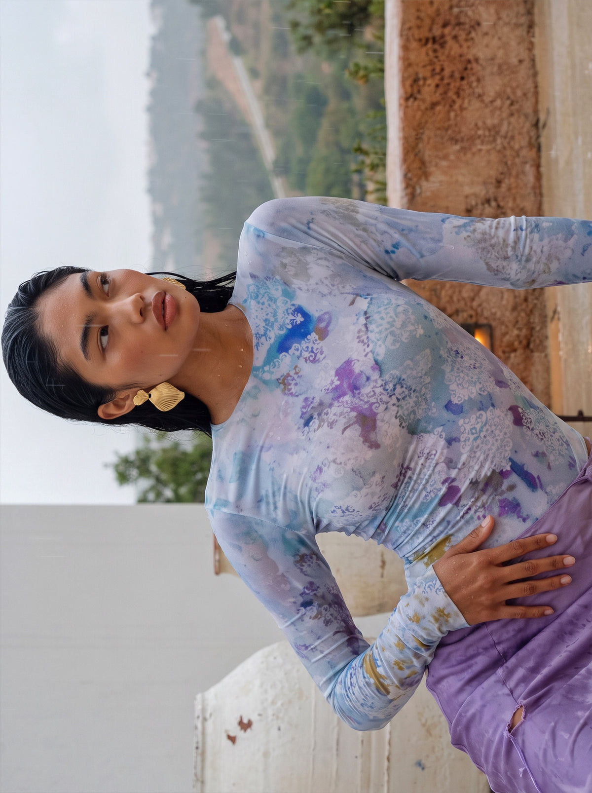 A woman wearing a long-sleeve fitted top with a blue, purple, and white watercolor-style print, paired with purple pants, posed outdoors against a rustic stone wall and distant landscape.