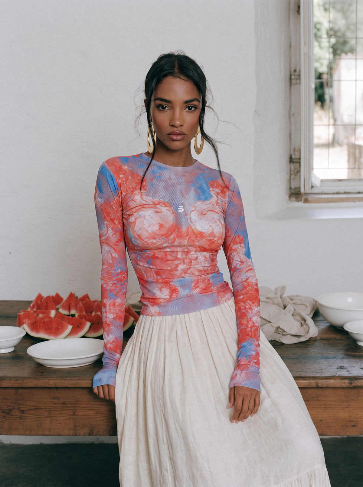 A woman stands indoors wearing a fitted long-sleeve top with a coral-red, blue, and lavender abstract print, paired with a cream pleated skirt. She is posed beside a wooden table with plates and watermelon slices against a white wall and window.