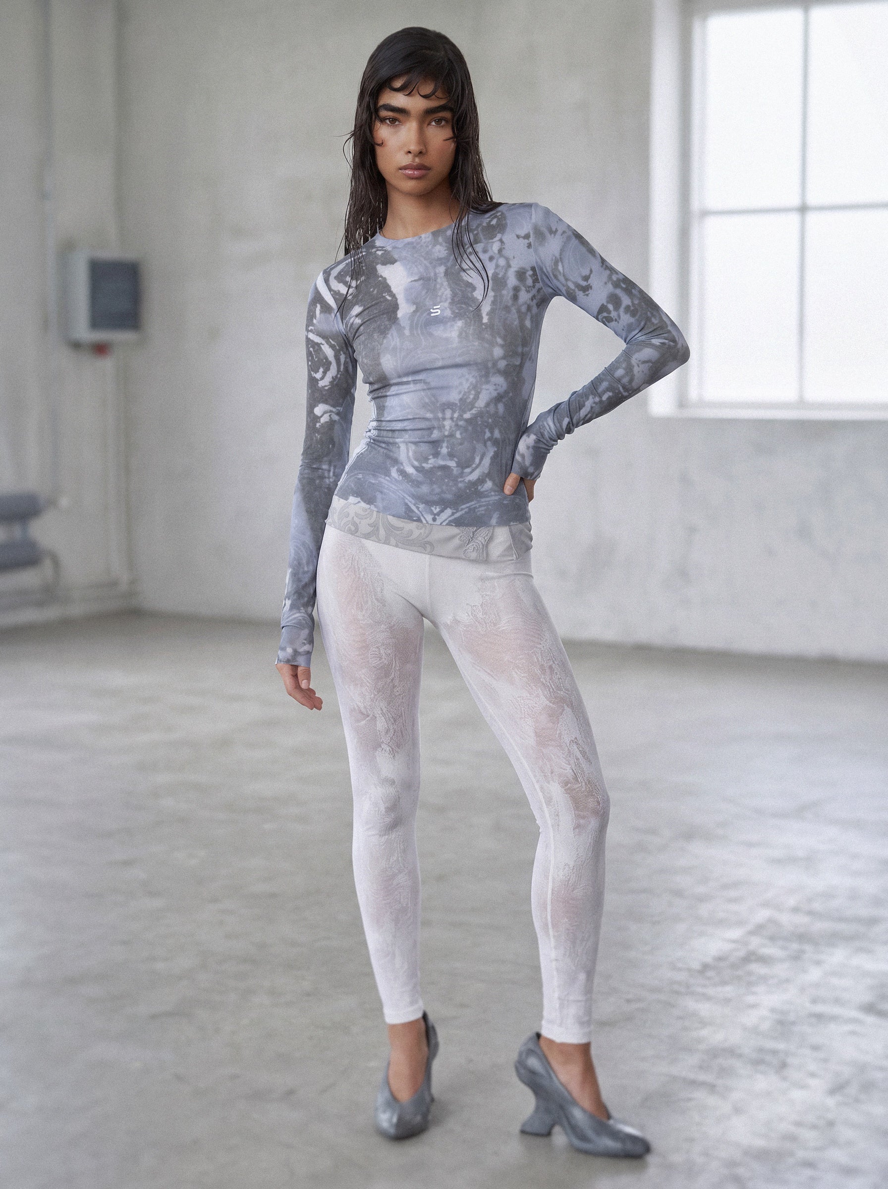 Woman wearing a patterned long-sleeve top and sheer white leggings in a minimalistic room.