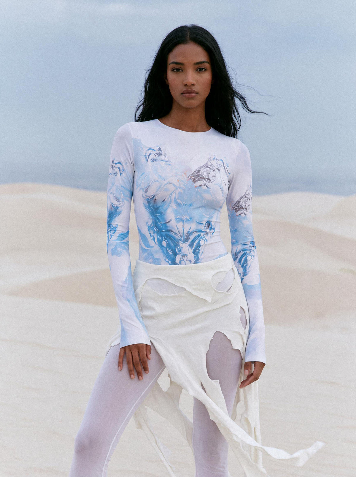 A model stands in a desert setting wearing a white long-sleeve top with a blue and gray abstract print, paired with a white asymmetrical draped skirt and white leggings.
