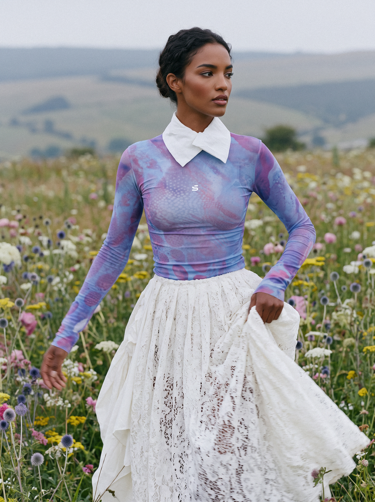 A woman stands in a field of wildflowers wearing a long-sleeve collared top with a purple and blue marbled pattern, paired with a white lace skirt.