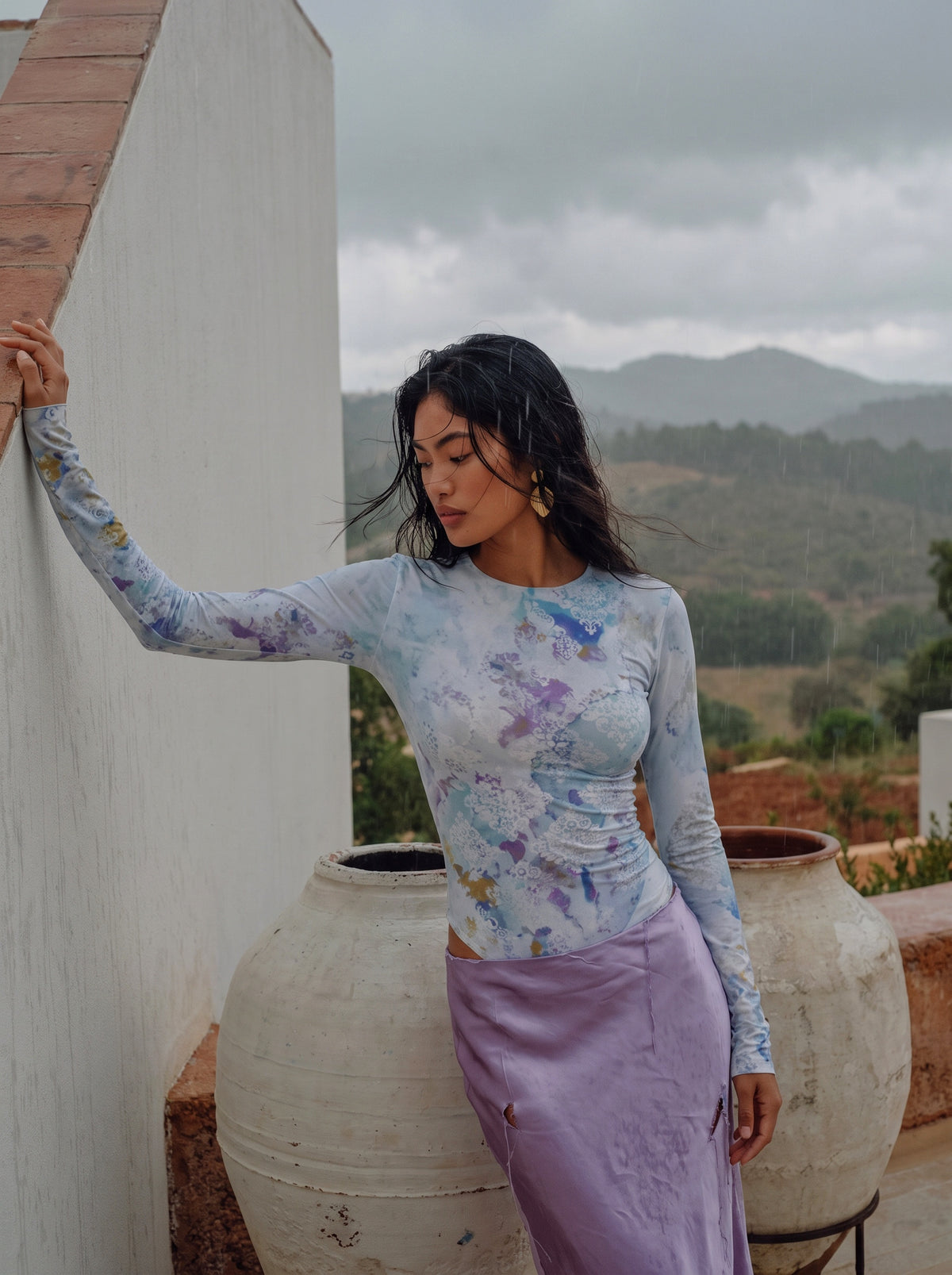 A woman stands outdoors against a white wall with large clay pots and a cloudy mountain landscape in the background, wearing a fitted long-sleeve top with a pastel blue, purple, and white abstract print paired with a lavender skirt.