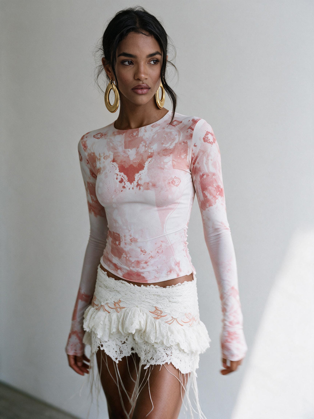 A woman modeling a fitted long-sleeve top with a pink and white tie-dye pattern, paired with a white fringed mini skirt, against a plain light gray wall.