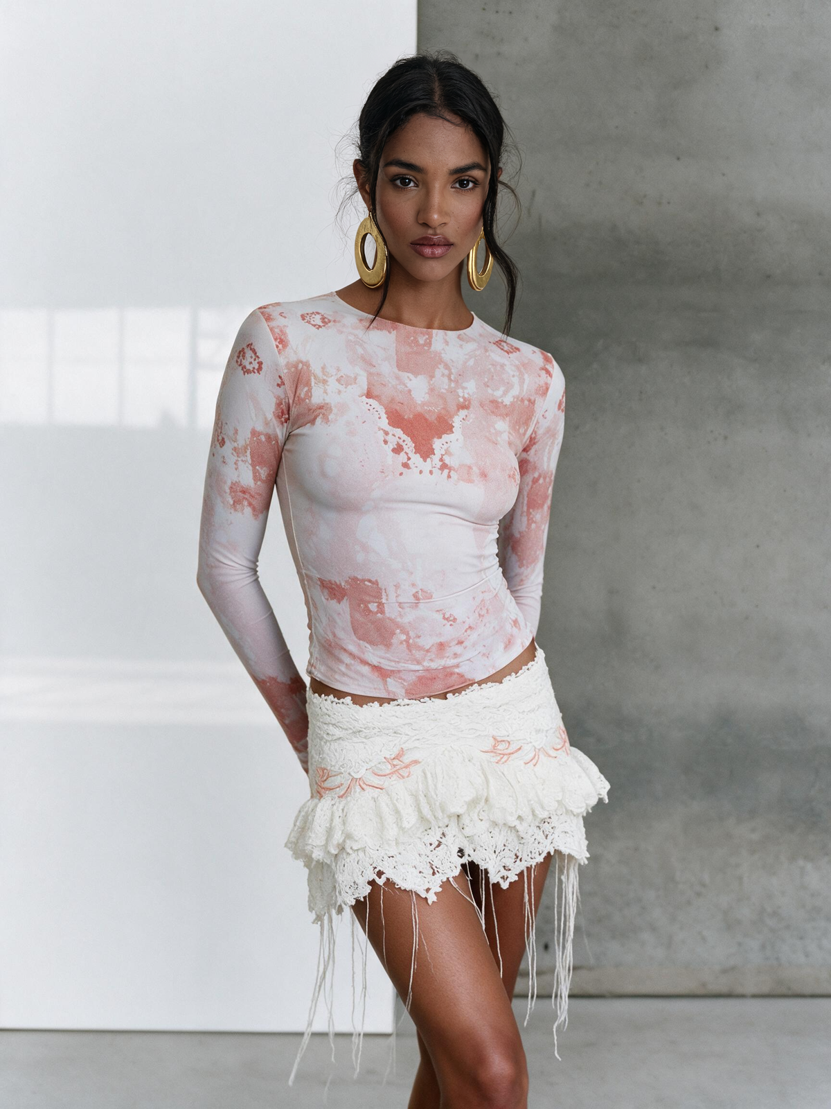 A woman modeling a long-sleeve fitted top with a pink and white abstract print, paired with a white fringed lace mini skirt, standing against a split white and gray studio background.