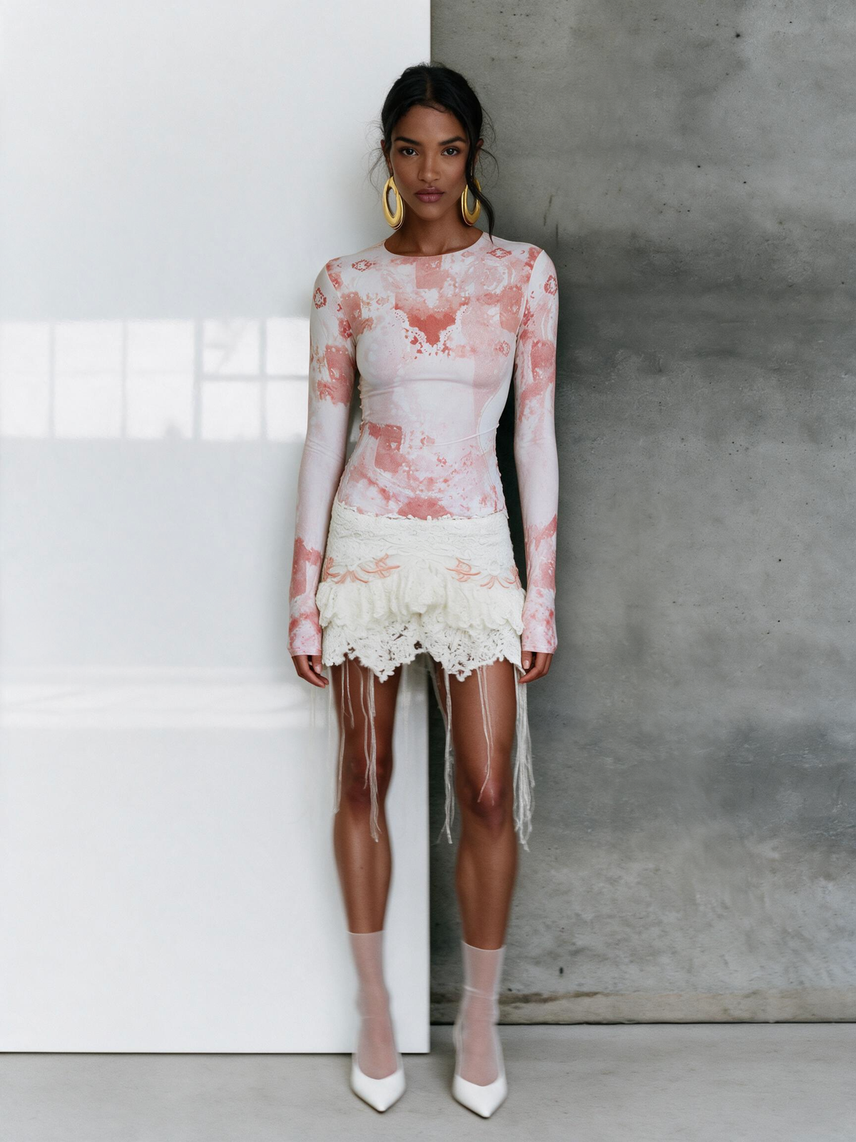 A model stands against a split white and gray wall wearing a sheer long-sleeve top and fringed mini skirt in a pink and white floral print, paired with sheer nude tights and white pointed heels.