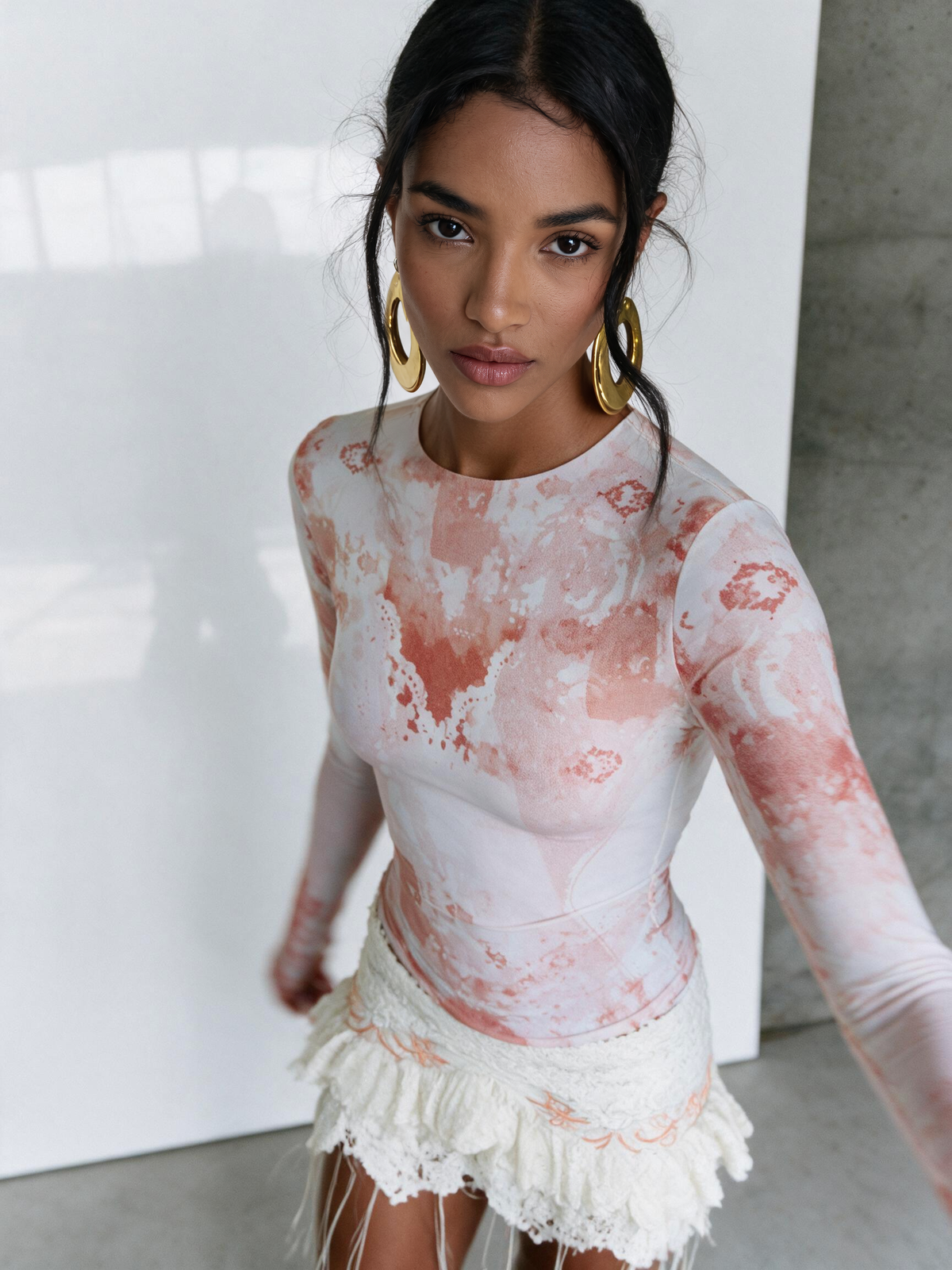 A woman wearing a fitted long-sleeve top with a pink and white tie-dye pattern, paired with a white fringed mini skirt and large gold hoop earrings, standing in a minimalist studio setting.
