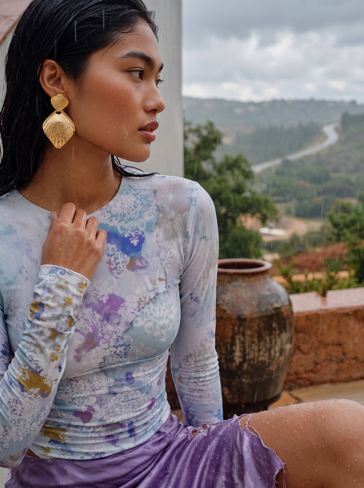 A woman wearing a long-sleeve tie-dye top in pastel blue, purple, and white, paired with a purple skirt, sitting outdoors with a scenic mountain background.