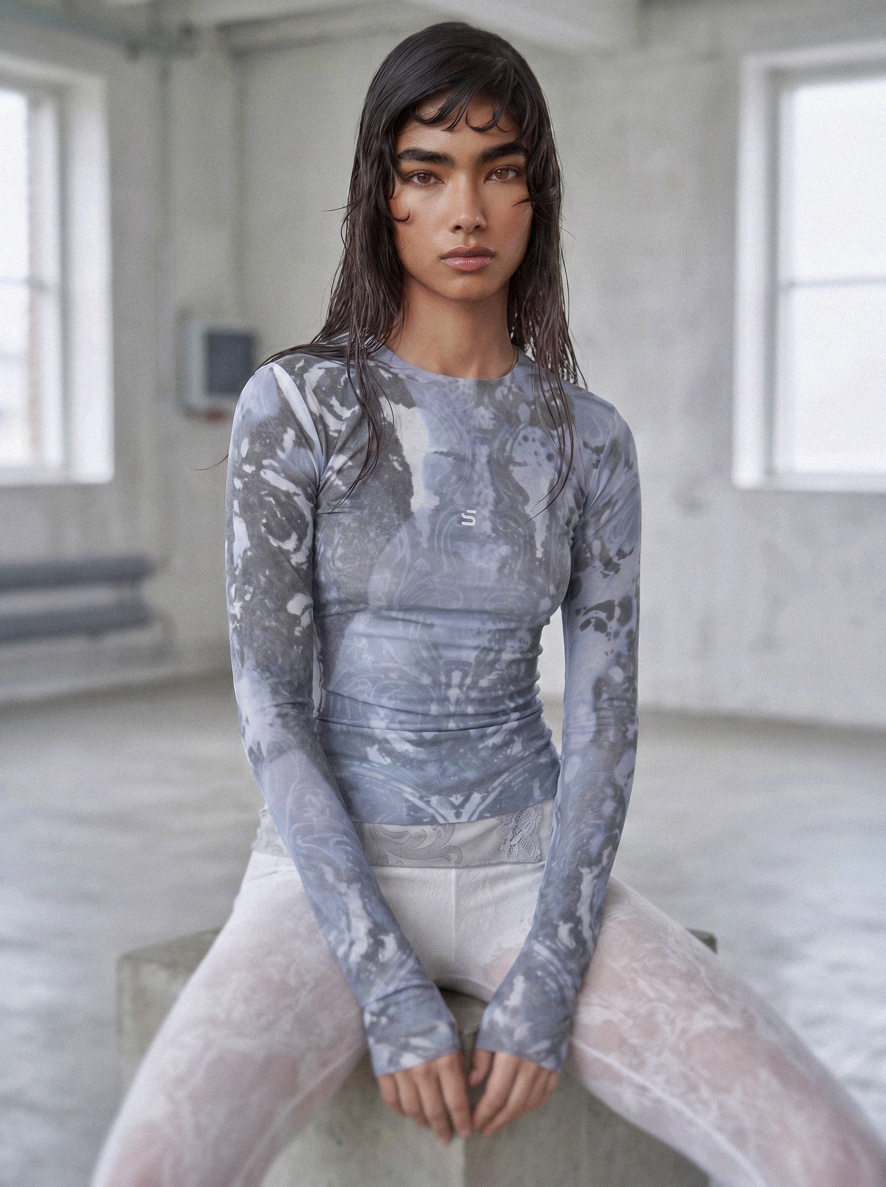 Person wearing a gray textured long-sleeve top and white pants in a minimalistic room.