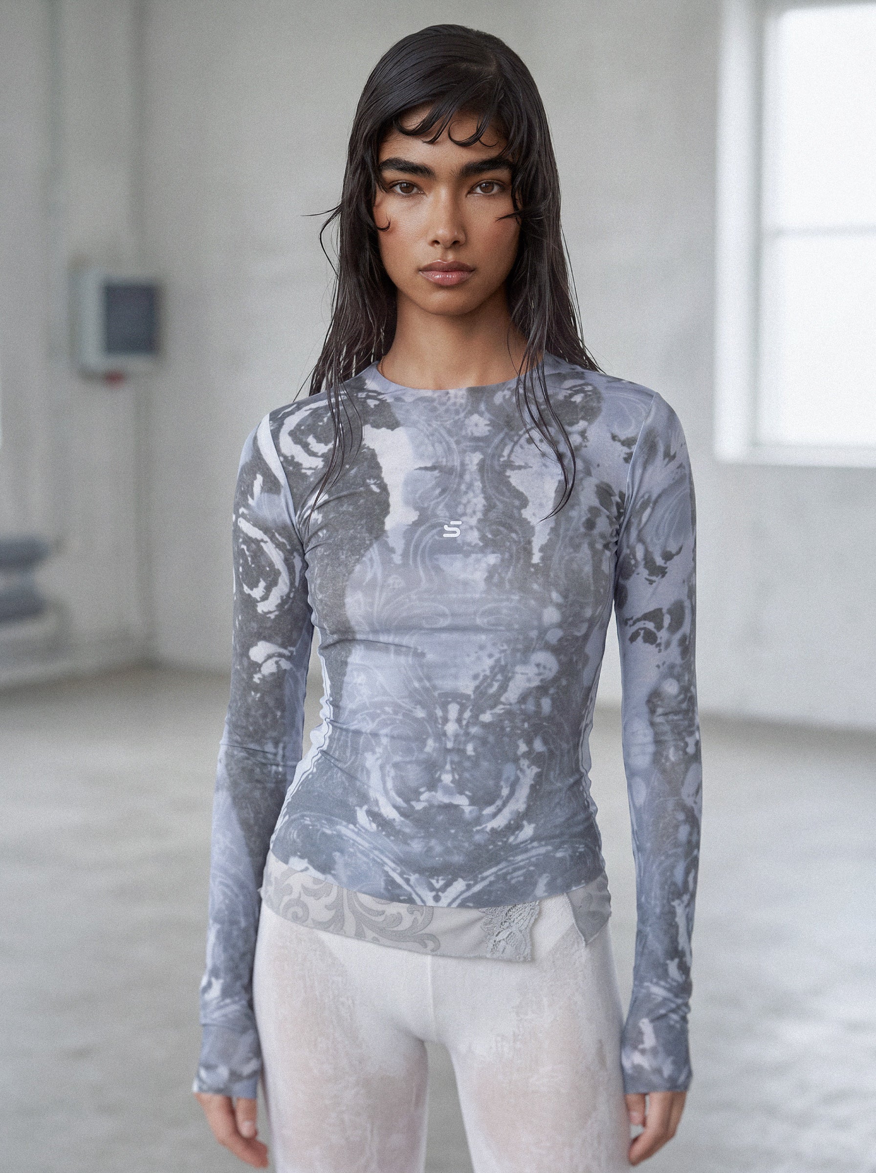 Model wearing a gray patterned long-sleeve top and white pants in a minimalistic setting.