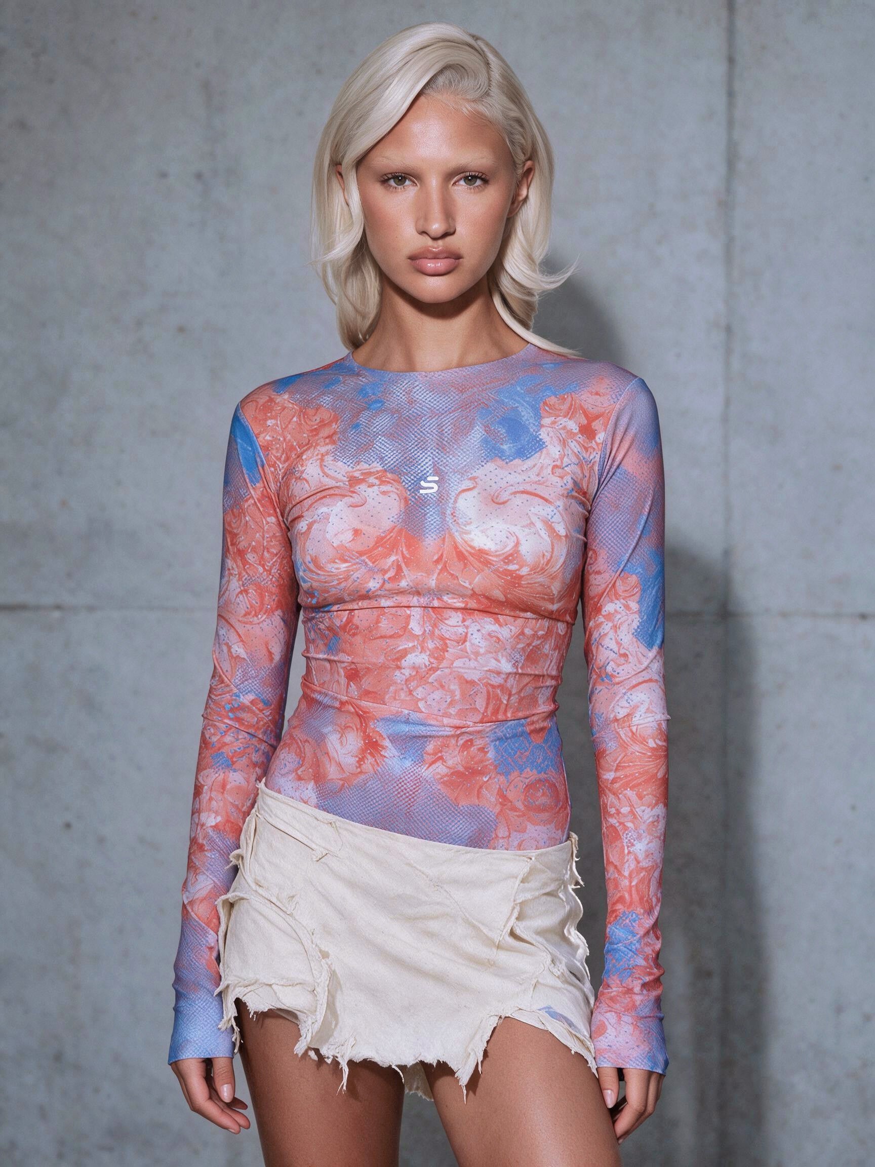 A blonde model wearing a long-sleeve fitted top with a pink, blue, and white abstract floral print, paired with a distressed cream mini skirt, standing against a textured gray concrete wall.